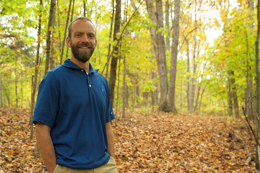 Blaine Adamson stands smiling in a sunny deciduous forest.